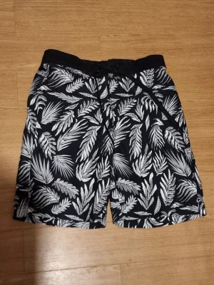 Svarta shorts med vit bladmönster - INSTABOX!!! Snygga svarta shorts med vitt bladmönster och snörning i midjan. Perfekta för sommaren med sin avslappnade passform och elastisk midja. Materialet känns lätt och bekvämt, perfekt för varma dagar.