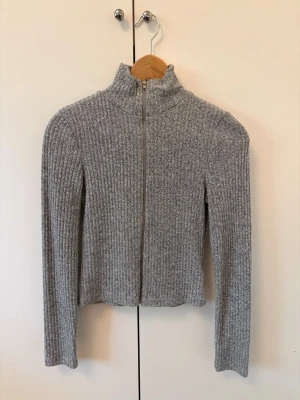 Grå ribbad ziptröja med hög krage - Säljer en stilren grå ribbad tröja med dragkedja framtill och hög krage. Tröjan har lång ärm och är tillverkad i ett mjukt, stretchigt material som sitter snyggt på kroppen. Perfekt för lager på lager och enkel att matcha.