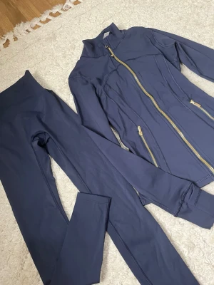 Marinblå  träningsset Lululemon  - Snyggt mörkblått träningsset med långärmad jacka och matchande tights. Jackan har guldfärgade dragkedjor framtill och vid fickorna samt figurnära passform. Materialet är stretchigt och passar perfekt för gym eller löpning.