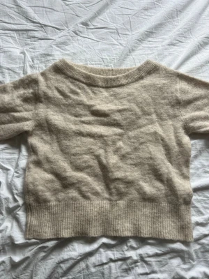 Beige stickad tröja med kort ärm - Mysig beige stickad tröja med korta ärmar och bred ribbad mudd vid nederkant och ärmslut. Tröjan har en rund halsringning och är tillverkad i ett mjukt, ulligt material som känns lyxigt och skönt mot huden.