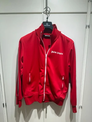 Röd track jacket från Palm Angels - Röd track jacket från Palm Angels med vit dragkedja och vita detaljer längs axlarna. Tröjan har hög krage, två fickor med dragkedja och logga tryckt på bröstet. Perfekt för en sportig och trendig look. Pris är diskuterbart!