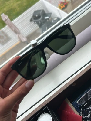 Svarta Ray-Ban solglasögon - Snygga svarta solglasögon från Ray-Ban med klassisk, fyrkantig form och mörka glas. Bågarna är i plast och har Ray-Ban-loggan på sidan. Perfekta för soliga dagar och ger en cool vibe till din outfit.saknar en spik så det bör fixas annars kan en av sidorna ramla av 