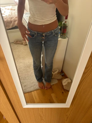 Ljusblå bootcut jeans med knappar - Säljer ett par ljusblå bootcut jeans med tre synliga knappar framtill och klassiska fickor både fram och bak. Jeansen har en lätt tvättad look och raka ben. Perfekta för en avslappnad och trendig stil.