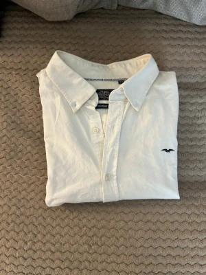 Vit skjorta med broderad logga - Klassisk vit skjorta med krage och knappar framtill. Skjortan har en liten broderad logga på bröstet och är tillverkad i ett mjukt bomullstyg. Perfekt till jeans eller chinos för en clean look. Jag tror att det passar för storlek L