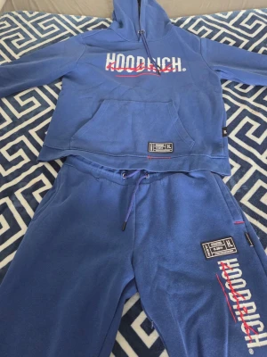 Blå HOODRICH loungewear set - Snyggt blått loungewear set från HOODRICH med hoodie och matchande byxor. Hoodien har stor magficka och tryckt logga framtill, medan byxorna har dragsko i midjan och HOODRICH-tryck på benet. Båda plaggen har detaljerade etiketter och är gjorda i mjukt material, perfekt för chill dagar.