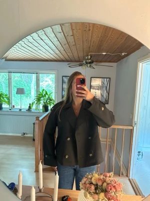 Svart kort trenchcoat med silvriga knappar - Stilren svart trenchcoat i kort modell med bred krage och dubbelknäppning med bruna knappar. Jackan är endast använd en gång. Passar även M. 