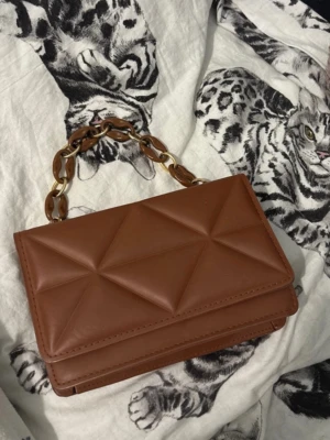 Brun quiltad handväska med kedjehandtag - Snygg brun handväska i quiltad design med geometriska mönster. Väskan har ett kort handtag i plastkedja med inslag av guld och brunt. Tillverkad i konstläder och har en stilren, rektangulär form som ger en modern vibe.