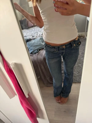 Blå low waist bootcut jeans - Säljer ett par blå low waist jeans med bootcut-modell. Jeansen har dubbla knappar fram, dekorativa fickor bak med lock och guldknappar samt klassisk femficksdesign. Perfekta för dig som gillar en retro och avslappnad stil.