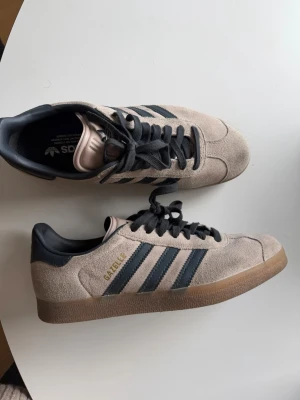 Adidas Gazelle beige sneakers - Säljer ett par klassiska Adidas Gazelle sneakers i beige mocka med mörkblå detaljer. Endast använt ett fåtal gånger. Storlek 41. Nypris 1600.