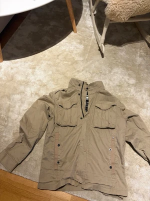 Beige vindjacka från G-Star RAW - Snygg beige vindjacka från G-Star RAW med hög krage och fyra framfickor med lock. Jackan har dragkedja och tryckknappar framtill samt RAW-logga på bröstet. Perfekt för dig som gillar streetwear och funktionell stil.