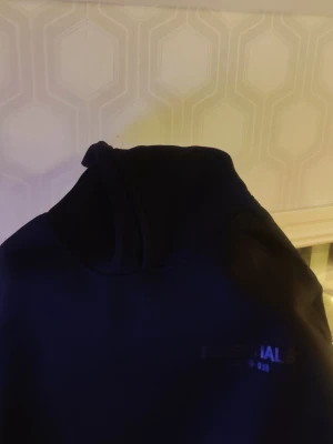 Svart Essentials Fear of God hoodie - Säljer en svart hoodie från Essentials Fear of God med stor logga tryckt på bröstet. Hoodien har klassisk huva och är gjord i mjukt material som passar perfekt för en chill och avslappnad stil.