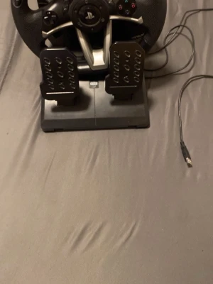 Ratt och pedaler till PlayStation - Säljer en svart racingratt med pedaler till PlayStation. Ratten har PlayStation-knappar, D-pad och två pedaler för gas och broms. Kopplas in med USB och har en sportig design i plast och metall-look. Perfekt för bilspel och racingupplevelser hemma. Funkar för ps4 och ps3