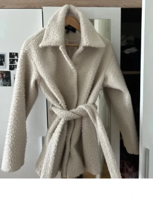 Beige teddyjacka med knytskärp - Supermjuk och fluffig teddyjacka i beige med stor krage och knytskärp i midjan. Jackan har långa ärmar och stängs med tryckknappar framtill. Perfekt för att hålla sig varm och snygg under kyliga dagar.