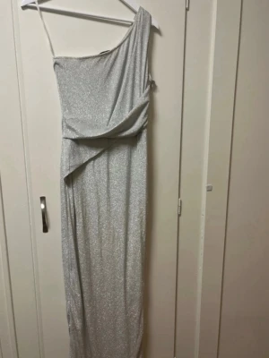Silverglittrig oneshoulder långklänning - Snygg silverfärgad långklänning med glittrigt tyg och oneshoulder-design. Klänningen har en asymmetrisk axel och ett draperat parti framtill som ger en cool och elegant look. Perfekt för dig som vill sticka ut på festen. Använd enbart i 3h. Storlek 10 
