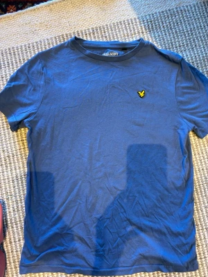 Blå t-shirt från Lyle & Scott - Säljer en blå t-shirt från Lyle & Scott med klassisk rund hals och korta ärmar. På bröstet sitter den ikoniska gula loggan. Perfekt till jeans eller shorts för en enkel och snygg look.