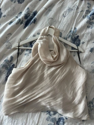 Champagnefärgad halterneck linnetopp - Säljer en snygg champagnefärgad halterneck topp i ett glansigt, tunt material. Toppen har en draperad halsringning och är ärmlös med en elegant, flowy passform. Perfekt för dig som vill ha en trendig och stilren look.