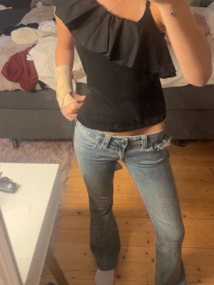 Svart omlottopp med volang - Säljer en svart topp med omlottkänsla och stor volangdetalj över ena axeln. Toppen är ärmlös och har en asymmetrisk design med en axelbandslös sida. Perfekt för dig som vill sticka ut lite extra. Lappen är borta men tror den är från Zara eller något liknande.