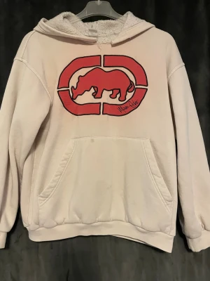 Hoddie - Ljusrosa hoodie med stor röd noshörningslogga på bröstet. Mjuk insida och huva med teddyfoder. Praktisk magficka och ribbade muddar vid ärmslut och nederkant. Perfekt för en avslappnad stil.