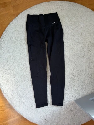 Svarta ribbade leggings Aim'n - Säljer ett par svarta ribbade leggings från Aim'n med hög midja och stretchigt material. De har en tight passform och diskret metallogo framtill i midjan. Perfekta för både träning och chill. Snyggt ribbat tyg som ger en trendig look.