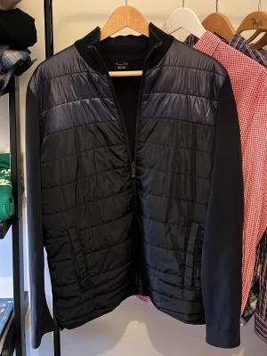Svart quiltad jacka Massimo Dutti - Snygg svart quiltad jacka från Massimo Dutti med lång ärm och dragkedja framtill. Jackan har en stilren design med diskreta paneler och hög krage. Perfekt för dig som vill ha en clean look med lite edge. Modell 180 77kg som referens!