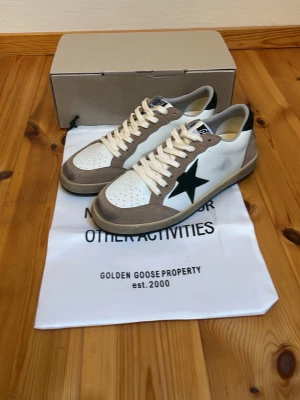 Golden Goose vita sneakers med stjärna - Snygga vita sneakers från Golden Goose med beige mockadetaljer och mörkgrön stjärna på sidan. Skorna har vita snören, perforerad tå och svart hälflik. Klassisk låg modell med retrovibbar och robust sula. Perfekta för dig som gillar streetstyle.