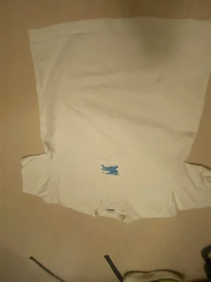 Vit Burberry  t-shirt med blått broderi - Vit t-shirt med ett blått broderat motiv på bröstet som föreställer en ryttare på häst. Klassisk passform och korta ärmar. Perfekt till jeans eller shorts för en enkel och stilren look.
