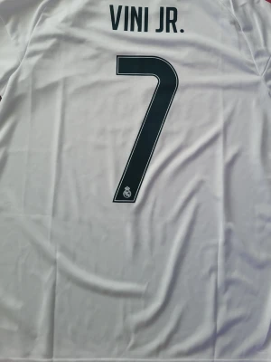 Real Madrid Vini Jr. matchtröja - Säljer en vit Real Madrid matchtröja med VINI JR. och nummer 7 på ryggen. Tröjan har mörkgröna tryck, röda ränder på axeln och klubbmärke på bröstet. Tillverkad i ett lätt och ventilerande material, perfekt för fotbollsträning eller match.