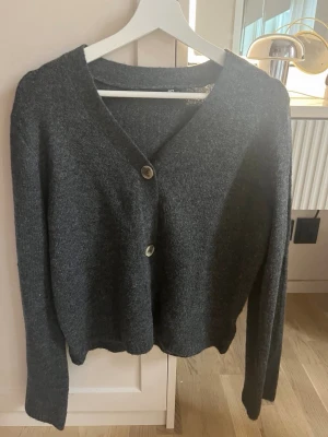Mörkgrå kofta från 157 - Mysig mörkgrå kofta från 157 med två stora knappar framtill. Koftan har v-ringning, lång ärm och är gjord i ett mjukt stickat material. Perfekt att slänga över en t-shirt eller linne för en chill look.