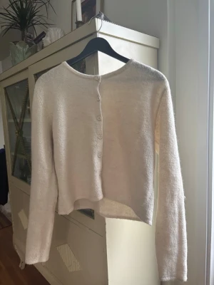 Krämvit mysig kofta med knappar - Mysig krämvit kofta med rund halsringning och knappar framtill. Koftan har lång ärm och är gjord i ett fluffigt, mjukt material som ger en avslappnad look. Perfekt att slänga över en topp för extra värme. Passar jättebra till ett kyligare höstväder! Storlek L men passar som en M.