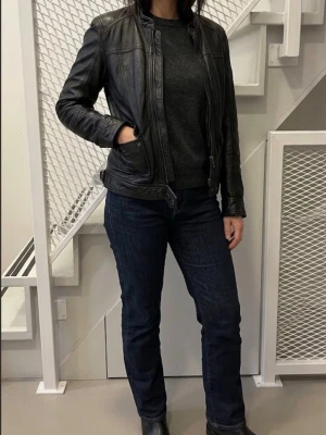 Svart skinnjacka med ståkrage - Snygg svart skinnjacka med ståkrage och dragkedja framtill. Jackan har två fickor med lock och knapp, samt detaljerade sömmar för en cool look. Passar perfekt till jeans och boots för en avslappnad stil.