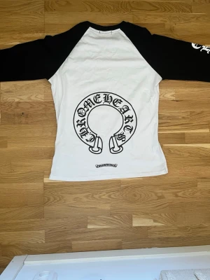 Chrome Hearts svartvit långärmad topp - Säljer en svartvit långärmad topp från Chrome Hearts med kontrastärmar och stora logotryck både fram och bak. Ärmarna har tryck längs hela sidan och halsen har också logga. Perfekt för dig som gillar streetwear och vill sticka ut.