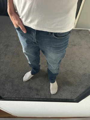 Jack & Jones jeans  - Säljer ett par klassiska blå jeans med rak passform och normal midja. Jeansen har fem fickor, bälteshällor och subtila slitningar framtill för en avslappnad look. Perfekta till vardagsstilen och passar till det mesta.