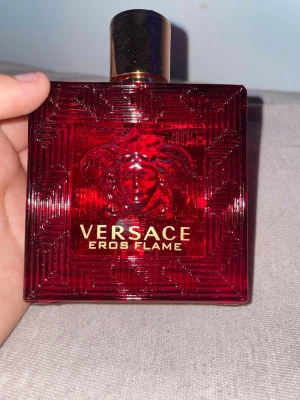 Versace Eros Flame Eau de Parfum - Versace Eros Flame Eau de Parfum i en lyxig röd glasflaska med gulddetaljer och Medusa-huvud i relief. Flaskan har en fyrkantig form och matchande röd kartong med guldfärgad text. Doften är intensiv och elegant, perfekt för dig som gillar exklusiva parfymer.