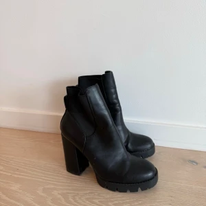 Svarta chunky stövletter med klack - Säljer ett par svarta stövletter med chunky klack och platåsula. Skorna har rundad tå och resår på sidorna för enkel påtagning. Tillverkade i ett slätt material som liknar skinn. Perfekta för dig som gillar en edgy och trendig look.