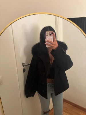 Gina tricot Svart pälskappa med stor huva - Säljer en svart päls kappa med stor fluffig huva och breda ärmar. Jackan har en oversized passform och stängs med knappar framtill. Perfekt för kalla dagar när du vill hålla dig varm och se snygg ut.