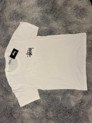 Vit Stüssy t-shirt med logga - Säljer en clean vit t-shirt från Stüssy med deras ikoniska svarta logga tryckt på bröstet. Klassisk rund hals och korta ärmar. Perfekt basic tee för dig som gillar streetwear och enkel stil.