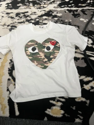 Vit t-shirt med camo-hjärta - Säljer en vit t-shirt med ett stort hjärta i camouflage-mönster och en liten röd detalj på bröstet. T-shirten har korta ärmar och en klassisk passform. Perfekt för dig som gillar streetwear och vill sticka ut lite extra.