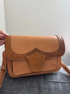 Brun axelväska i läderlook - Snygg axelväska i ljusbrunt och mörkbrunt konstläder med unik formad lockdetalj. Väskan har justerbar axelrem med metallspänne och dekorativa sömmar. Perfekt för dig som gillar retrostil och vill ha något som sticker ut. Mått 26x20x10 cm 