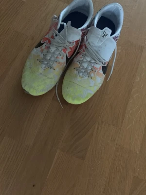 Färgglada Nike fotbollsskor - Säljer ett par Nike fotbollsskor med färgstarkt mönster i vitt, gult, rött och blått. Skorna har klassisk snörning, låg profil och svart Nike-logga på sidan. Perfekta för dig som vill sticka ut på planen!