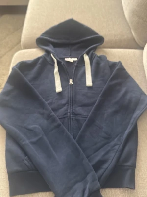 Mörkblå zip hoodie med vita snören - En mörkblå hoodie med dragkedja och vita snören i huvan. Klassisk känguruficka framtill och ribbade muddar vid ärmslut och nederkant. Perfekt för en avslappnad stil och enkel att matcha med jeans eller joggers.