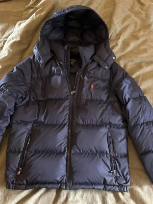 Mörkblå dunjacka från Polo Ralph Lauren - Snygg mörkblå dunjacka från Polo Ralph Lauren med huva och dragkedja framtill. Jackan har två sidofickor med dragkedja och en liten röd logga på bröstet. Perfekt för kalla vinterdagar tack vare det tjocka dunfodret. Använd under en sesong mycket bra skick. Stolek 18/20 xl motsvarar S.