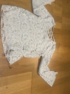 Vit spetsblus med omlott och v-ringning - Säljer en vit blus i spets med stora blommönster, v-ringning med korsade band framtill och omlottdetalj bak. Blusen har långa ärmar och är lätt transparent, perfekt för en trendig look.