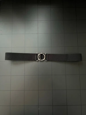 Svart elastiskt bälte med metallspänne - Snyggt svart bälte i elastiskt material med ett stilrent, runt metallspänne framtill. Perfekt för att ge en clean look till jeans eller klänning. Bältet är enkelt att justera och har en minimalistisk design.