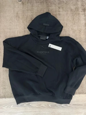  Essentials hoodie - Säljer en svart hoodie från Essentials Fear of God med diskret logga på bröstet. Hoodien har en klassisk huva, ribbade muddar och en relaxed fit. Tillverkad i mjukt material som känns skönt mot huden. Perfekt för dig som gillar streetwear och minimalistisk stil. Fint skick och sitter som en oversized m. Tags medföljer. 