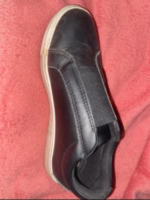 Svarta slip-on sneakers - Stilrena svarta slip-on sneakers med elastiskt band över vristen och vit sula. Skon har en enkel design utan snörning och är tillverkad i ett slätt material som ger en clean look. Perfekt för dig som gillar minimalistisk stil.