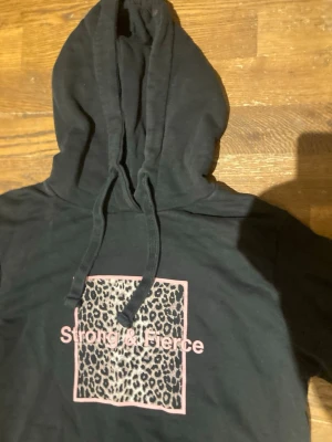 Svart hoodie med tryck från NA-KD, strl S - Svart hoodie utan någon ficka framtill, från NA-KD i storlek Small. På bröstet finns ett leopardmönstrat tryck i en rosa ram med texten Strong & Fierce i rosa. Finns några stycken fläckar på tröjan både på baksidan (se bild 5) och även framtill på huvan (se bild 3). Säljes pga för liten🤗