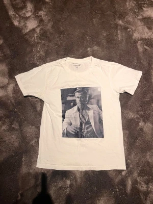 Vit t-shirt med tryck från Rovior - Vit t-shirt från Rovior med ett svartvitt tryck av en man i kostym på framsidan. Klassisk rund halsringning och korta ärmar. Perfekt för dig som gillar stilrena plagg med en twist. Xs 
