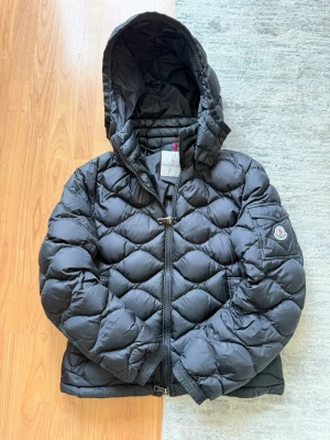 Moncler Jacka - Snygg svart dunjacka från Moncler ( Morandieres heter modellen ) med quiltat mönster och stor huva. Jackan har dragkedja framtill, ribbade muddar och en Moncler-logga på ärmen. Perfekt för kalla vinterdagar och riktigt varm tack vare dunfyllningen. Storlek 2 (S/M) 