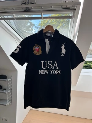 Svart Ralph Lauren pikétröja - Svart pikétröja med vit krage och detaljer, stor broderad logga på bröstet, USA och New York-tryck samt nummer 5 på ärmen och ryggen. Klassisk polokrage och korta ärmar. Perfekt för en sportig och stilren look. Ofta kallad ”Chief Keef” tröja. 