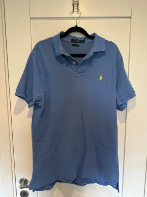Blå pikétröja från Polo Ralph Lauren - Säljer en blå pikétröja från Polo Ralph Lauren i slim fit-modell. Tröjan har korta ärmar, klassisk krage och den ikoniska gula loggan broderad på bröstet. Perfekt för en stilren och avslappnad look. Junior storlek large. Passar mig som brukar använda storlek medium i vuxen storlekar.